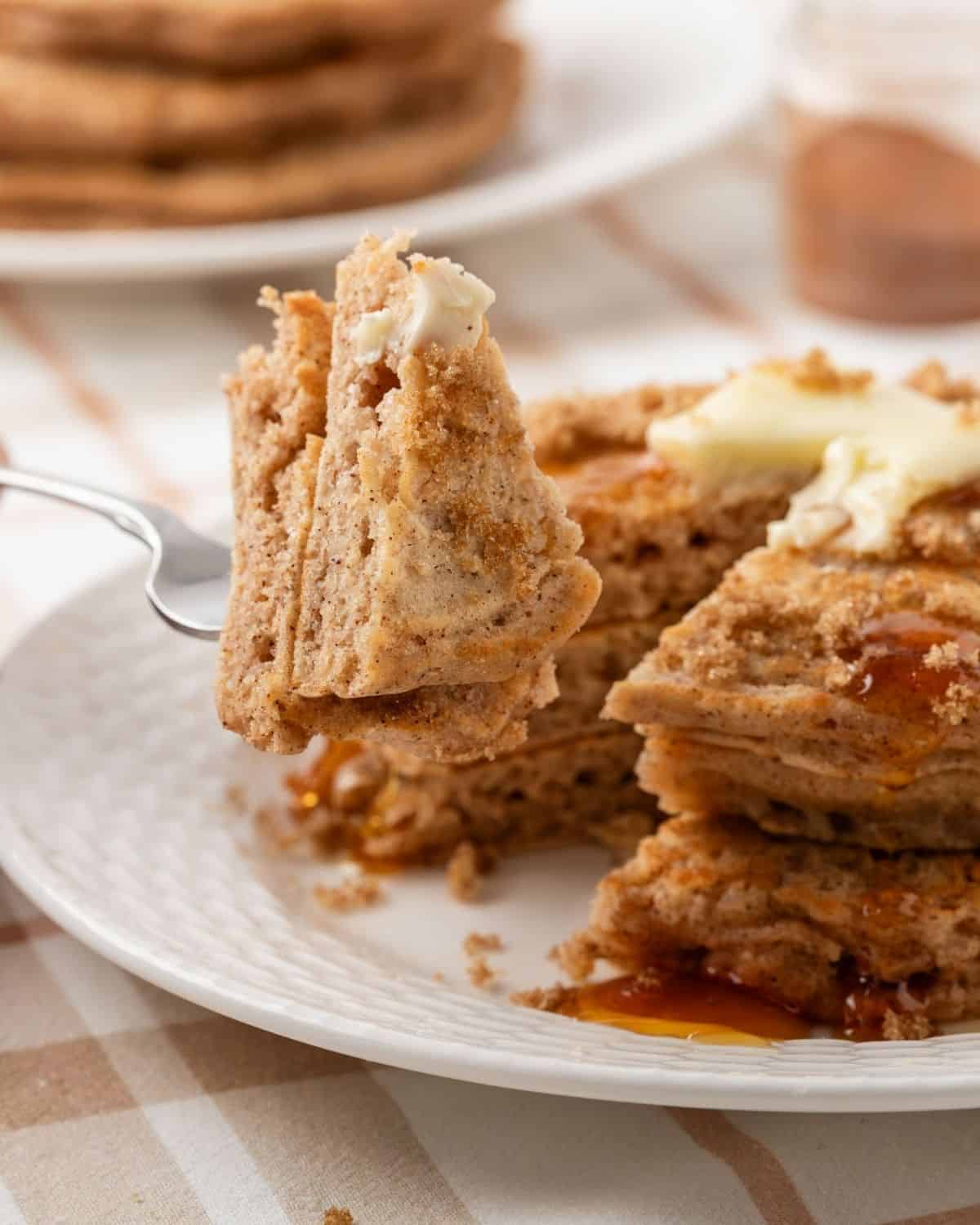 stack of cinnamon pancakes on a plate topped with a little butter, brown sugar, and pancake syrup and a piece pierced through a fork showing their fluffy texture