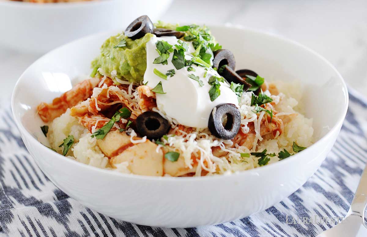 image: a white bowl filled with cauilflower rice, chicken, shredded cheese, guacamole, sour cream and sliced olives