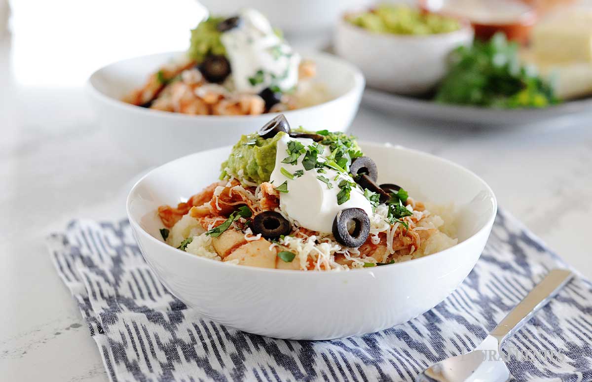 image: Two white bowls filled with cauilflower rice, chicken, shredded cheese, guacamole, sour cream and sliced olives