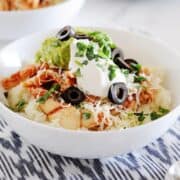 a white bowl filled with cauilflower rice, chicken, shredded cheese, guacamole, sour cream and sliced olives