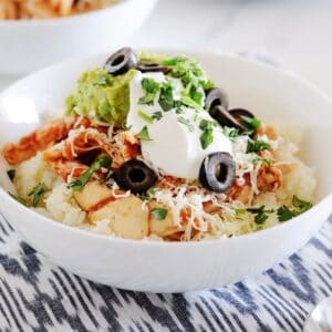 a white bowl filled with cauilflower rice, chicken, shredded cheese, guacamole, sour cream and sliced olives