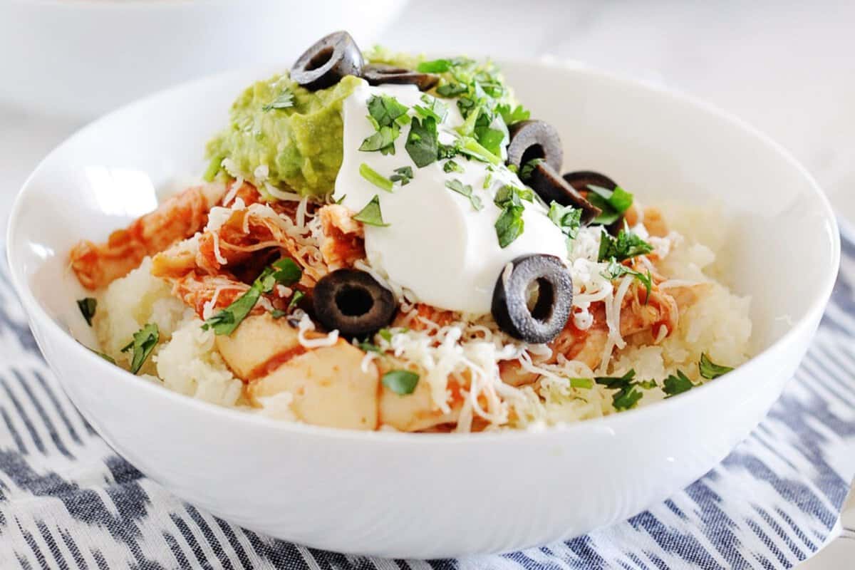 a white bowl filled with cauilflower rice, chicken, shredded cheese, guacamole, sour cream and sliced olives