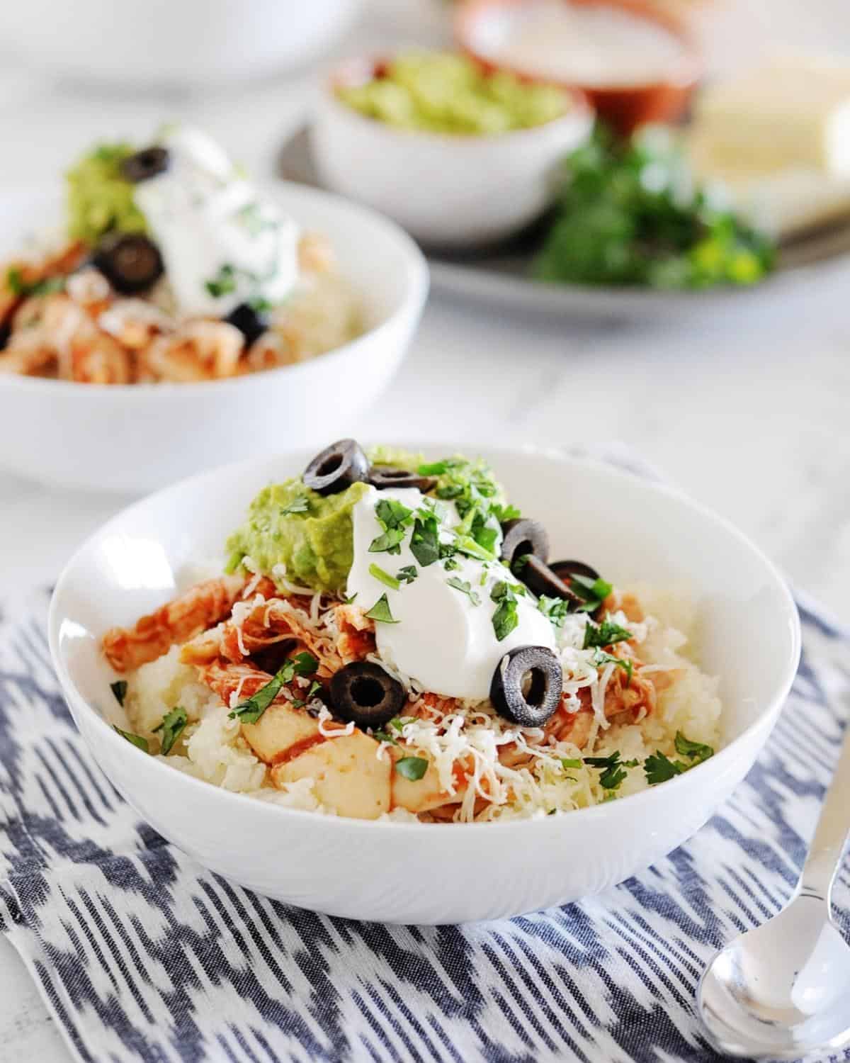 two white bowls filled with cauilflower rice, chicken, shredded cheese, guacamole, sour cream and sliced olives