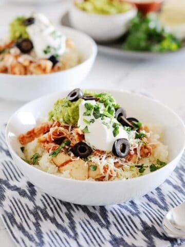 two white bowls filled with cauilflower rice, chicken, shredded cheese, guacamole, sour cream and sliced olives