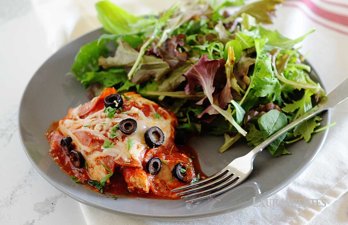 gray plate with pepperoni pizza chicken and large salad. Background of photo has skillet full of more pizza chicken.