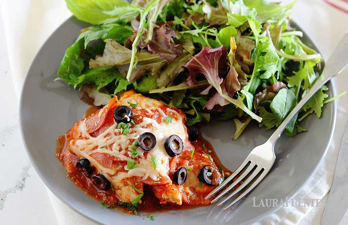 gray plate with pepperoni pizza chicken and large salad. Background of photo has skillet full of more pizza chicken.