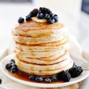 Stack of fluffy cassava flour pancakes on a plate. Topped with blackberries, blueberries, butter, and syrup.
