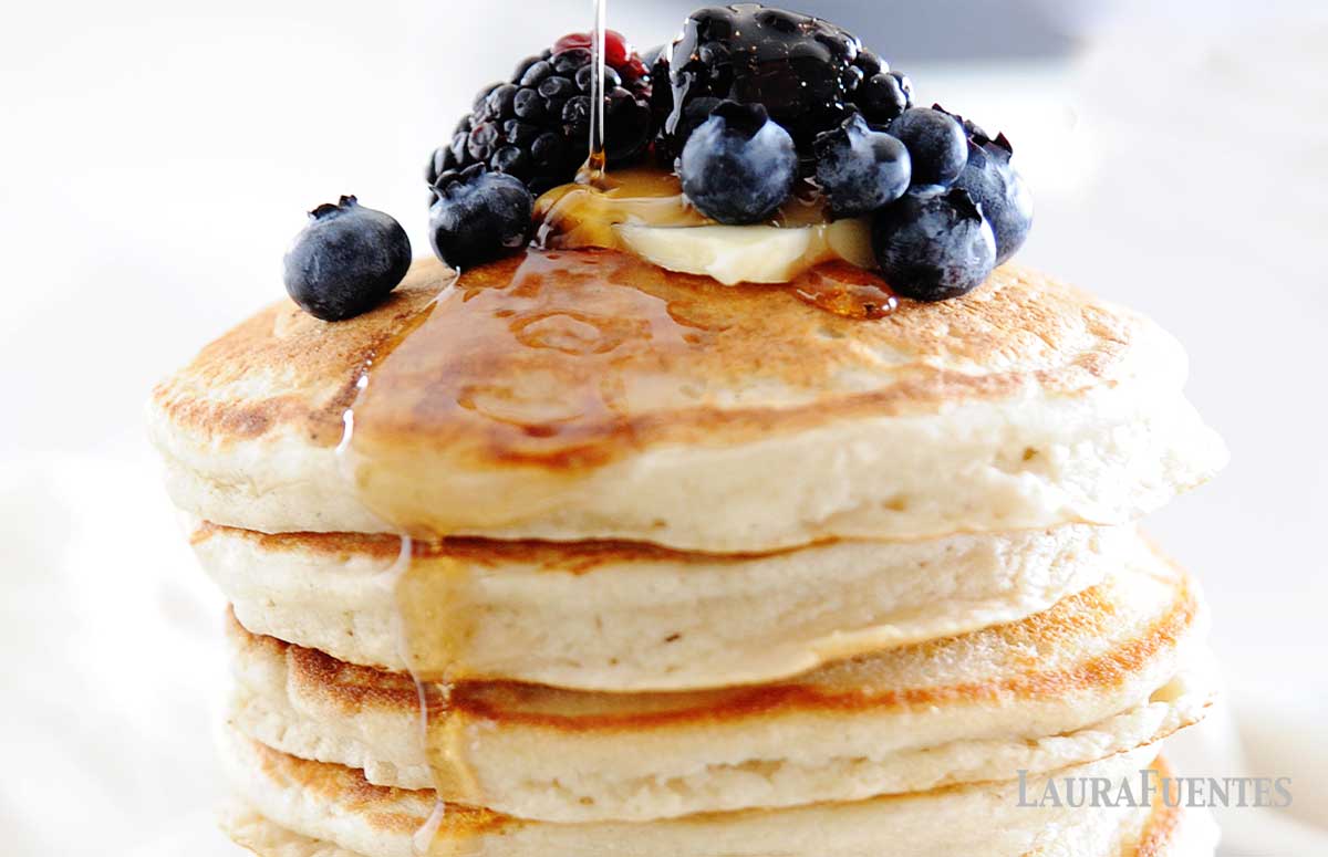 pouring syrup on a stack of cassava flour pancakes with blueberries and butter on top