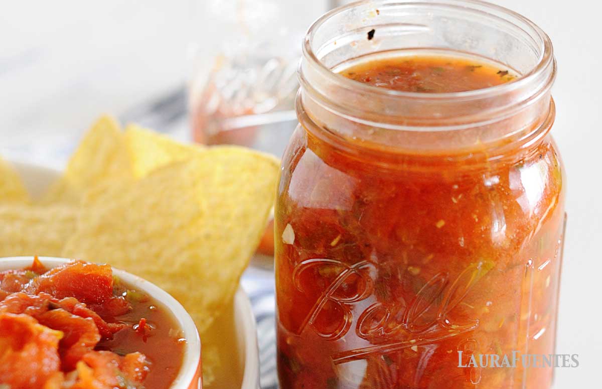 image: Open mason jar full of red salsa next to dish of corn chips and bowl of the same salsa