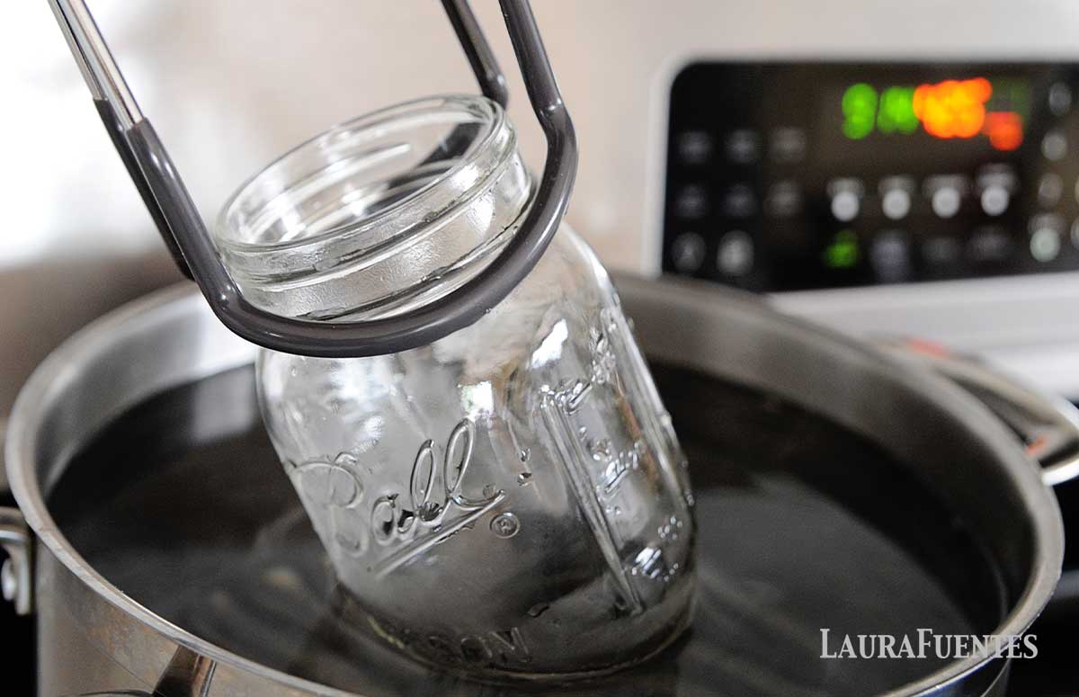 image: tongs lifting a canning jar out of a pot of hot water on a stove