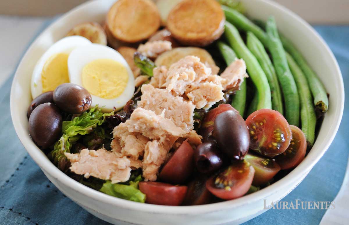 salmon nicoise salad topped with hard-boiled eggs, salmon, cherry tomatoes, green beans, potatoes, and olives in a white bowl.