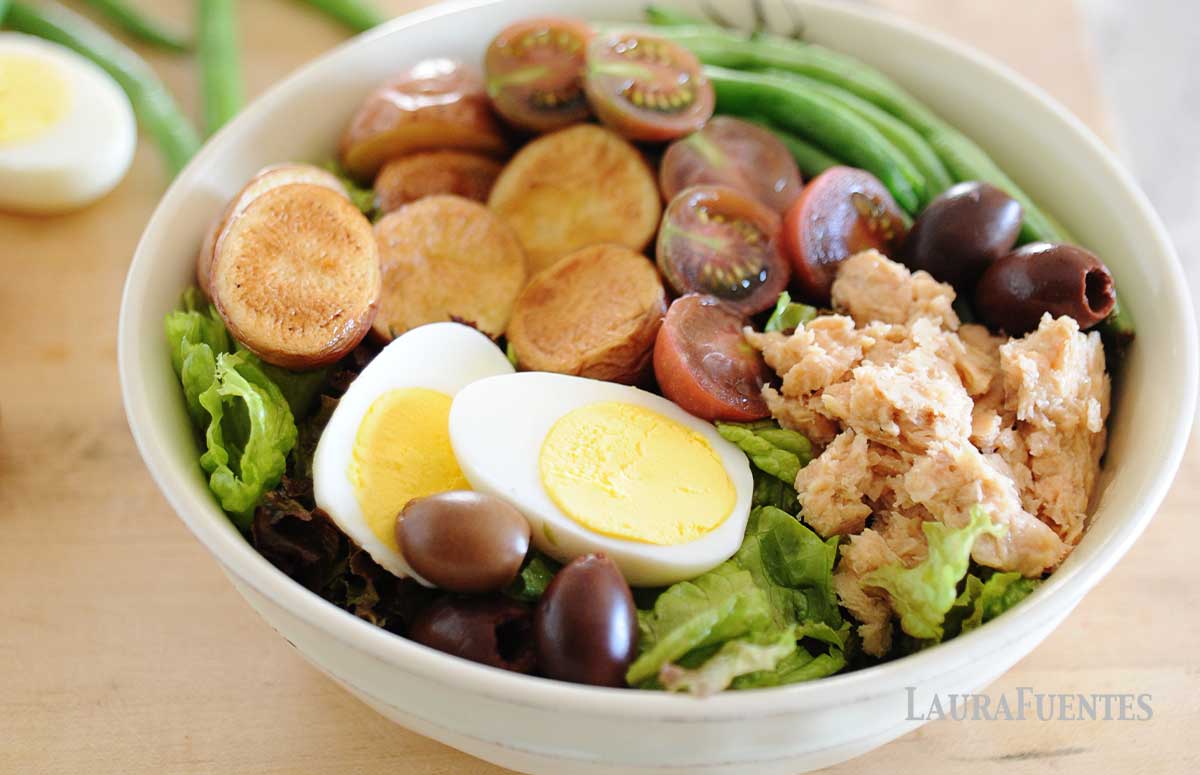 salmon nicoise salad topped with hard-boiled eggs, salmon, cherry tomatoes, green beans, potatoes, and olives in a white bowl.