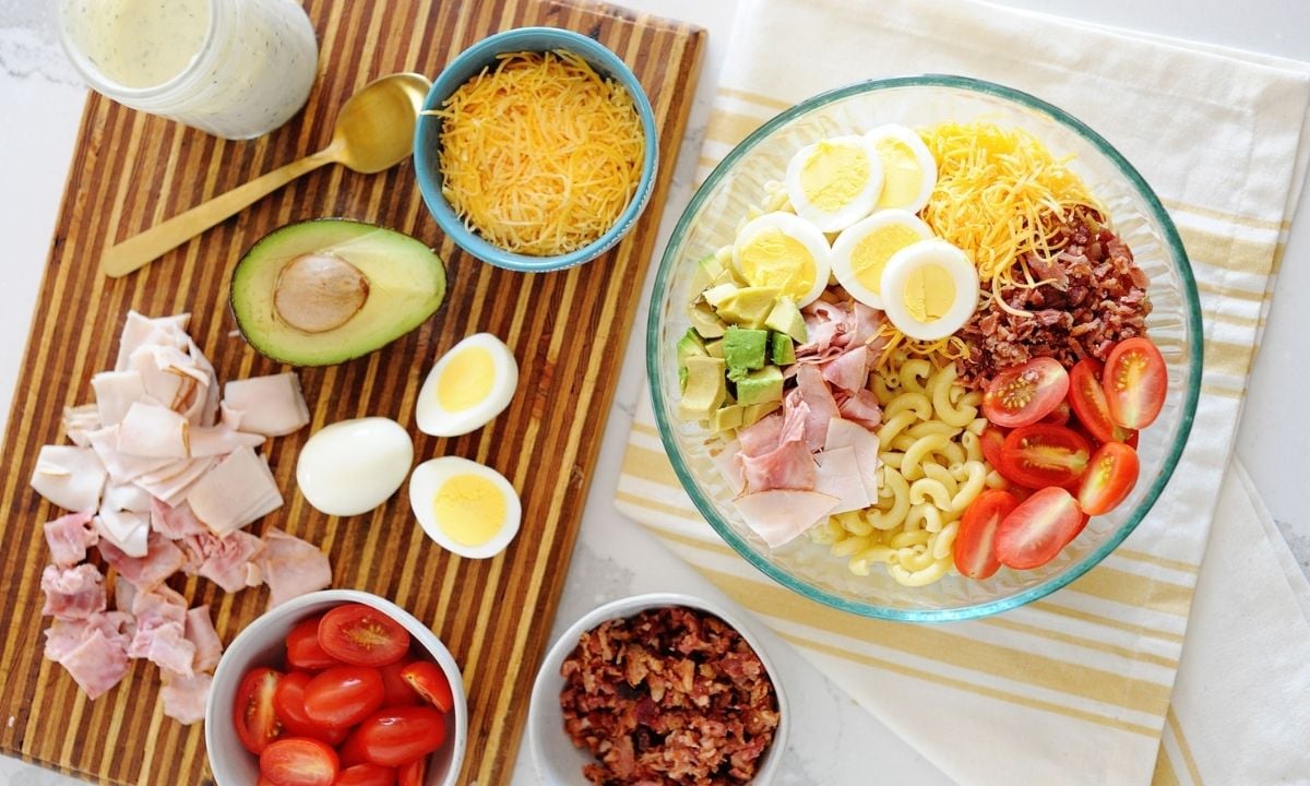 ingrediets for cobb pasta salad on a wooden board and a bowl: avocado, ham, bacon, hard boiled egg, tomatoes, cheese and dressing