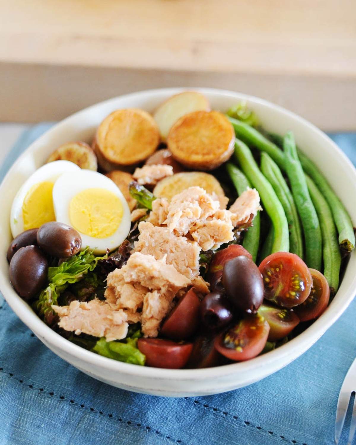 salmon nicoise salad topped with hard-boiled eggs, salmon, cherry tomatoes, green beans, potatoes, and olives in a white bowl.