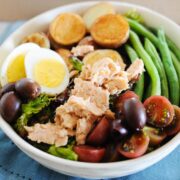 salmon nicoise salad topped with hard-boiled eggs, salmon, cherry tomatoes, green beans, potatoes, and olives in a white bowl.