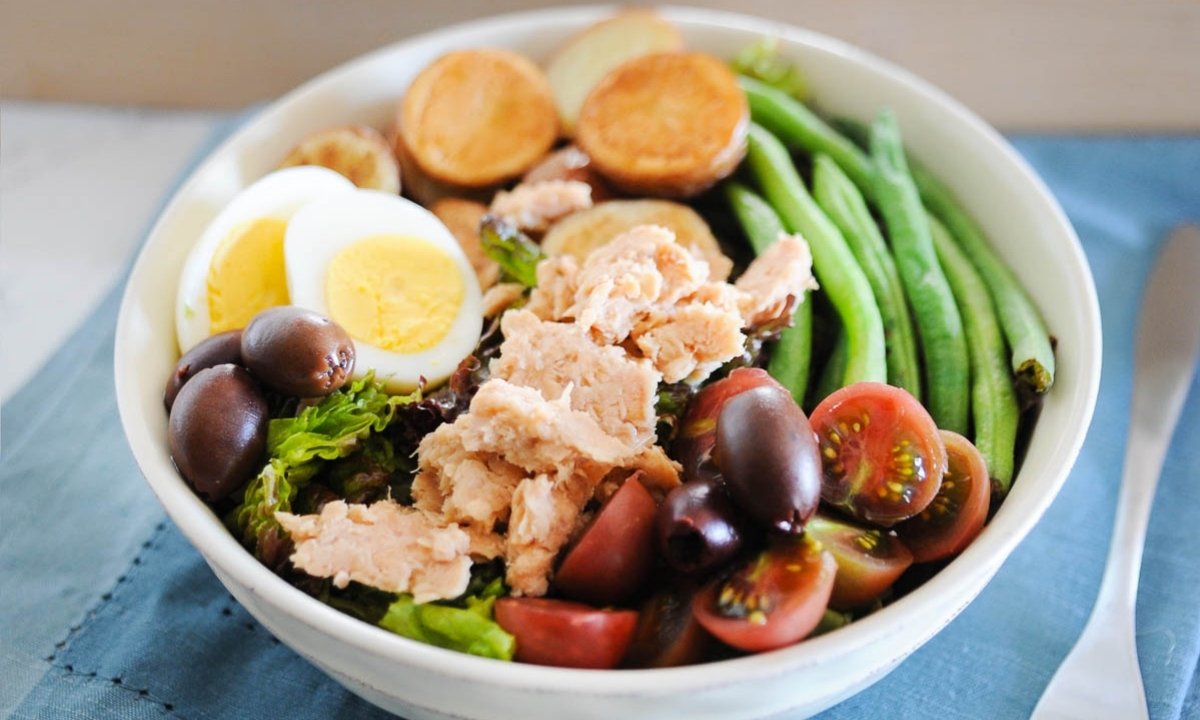 salmon nicoise salad topped with hard-boiled eggs, salmon, cherry tomatoes, green beans, potatoes, and olives in a white bowl.