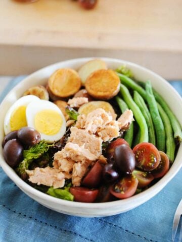 salmon nicoise salad topped with hard-boiled eggs, salmon, cherry tomatoes, green beans, potatoes, and olives in a white bowl.