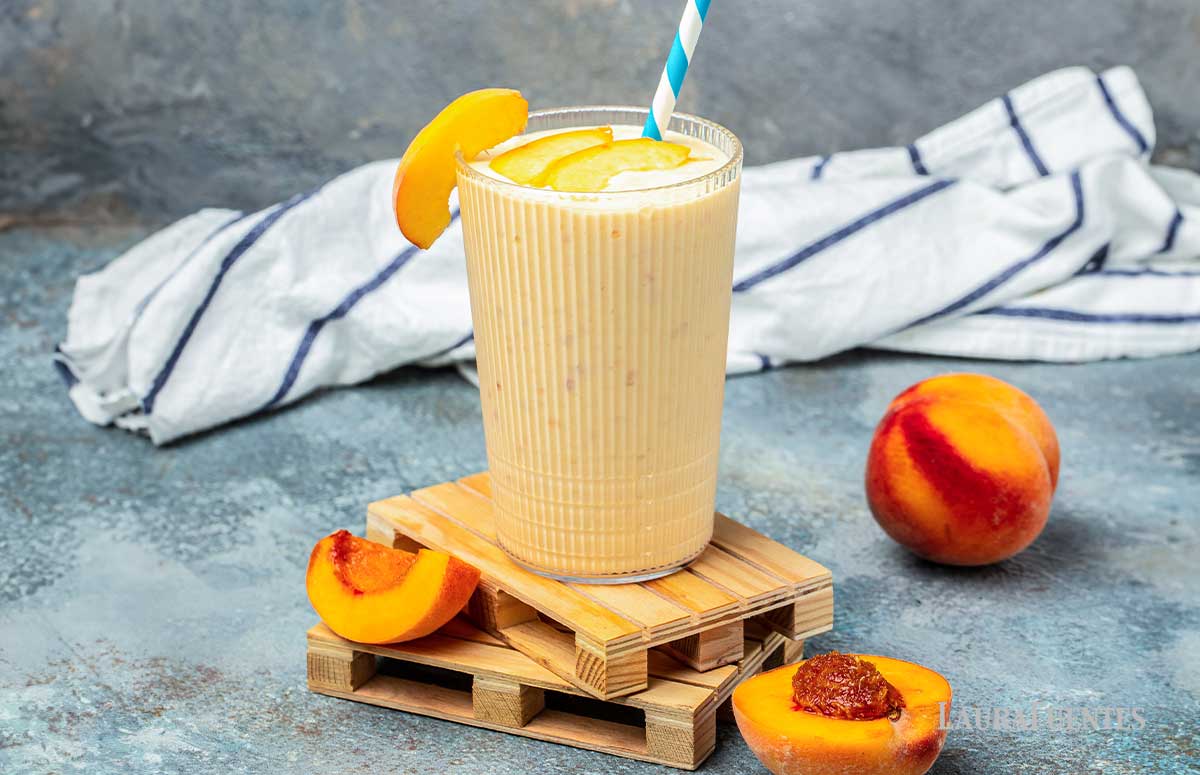a glass of peach cobbler smoothie with a dish towel behind and a few peaches around