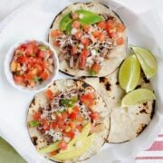 two street tacos next to a little bowl of pico de gallo and lime wedges