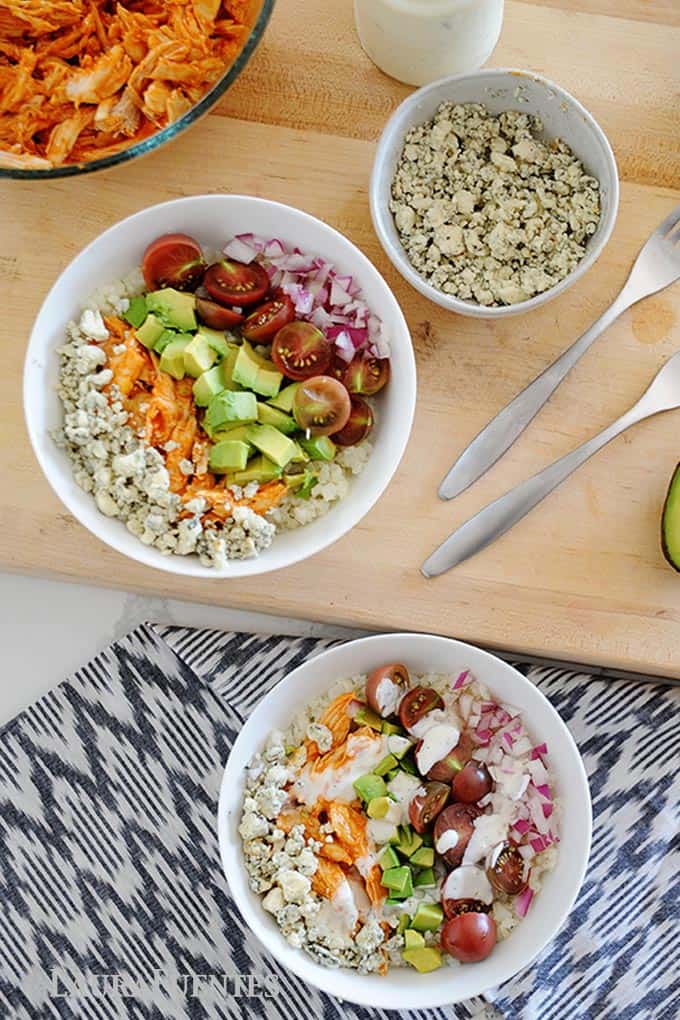 two white bowls of buffalo chicken, rice and vegetables. Bowls of ingredients like chicken and cheese to the side.