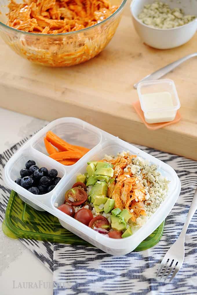 image: buffalo chicken, avocado cubes, halved tomatoes and cheese crumbles in a lunch container with carrot sticks and blueberries on the side.