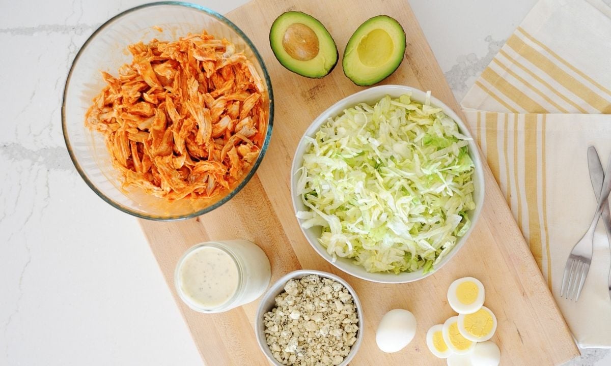 ingredients for buffalo chicken salad: bowl with buffalo chicken, shredded lettuce, crumbled cheese, a jar of dressing, avocado and hard boiled eggs