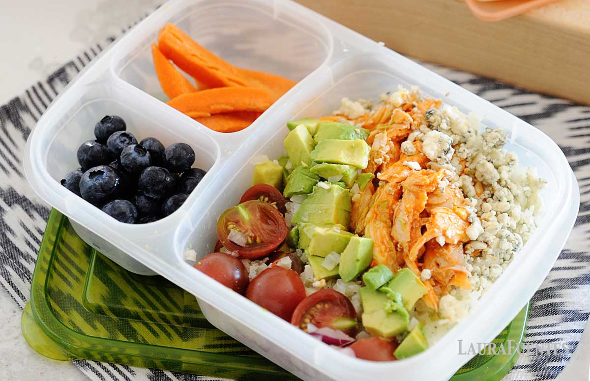 buffalo chicken, avocado cubes, halved tomatoes and cheese crumbles in a lunch container with carrot sticks and blueberries on the side