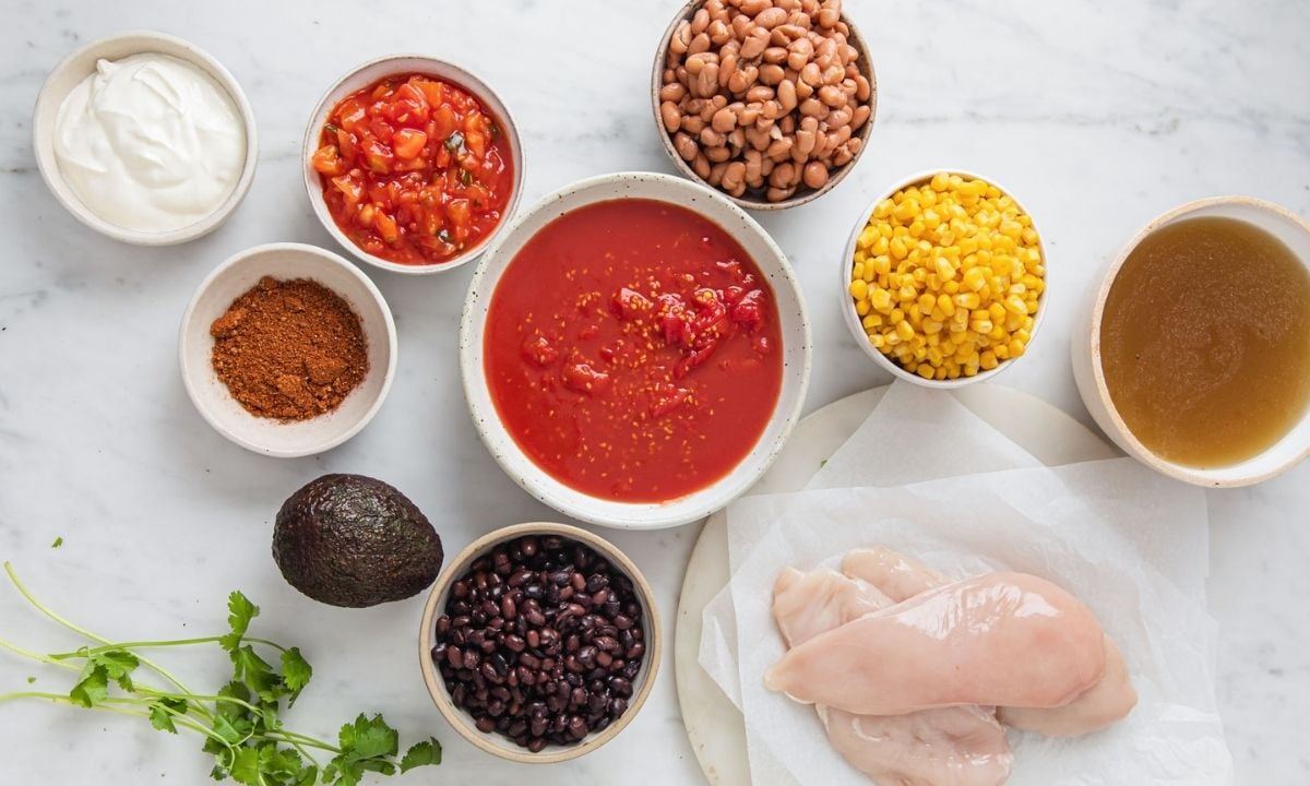 Slow cooker chicken taco soup ingredients with chicken breasts, diced tomatoes, black beans, pinto beans, corn, chicken broth, taco seasoning, salsa, avocado, sour cream, and cilantro.