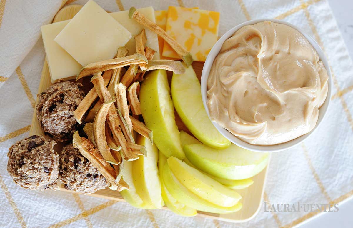 Apple slices, dried apples, cheese slices and energy bites on a board with a bowl of homemade caramel dip