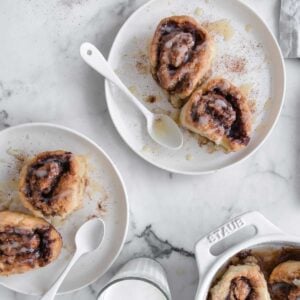 image: Two plates with two huge gluten-free cinnamon rolls on top. Next to a cup of milk and a dish of cinnamon rolls.