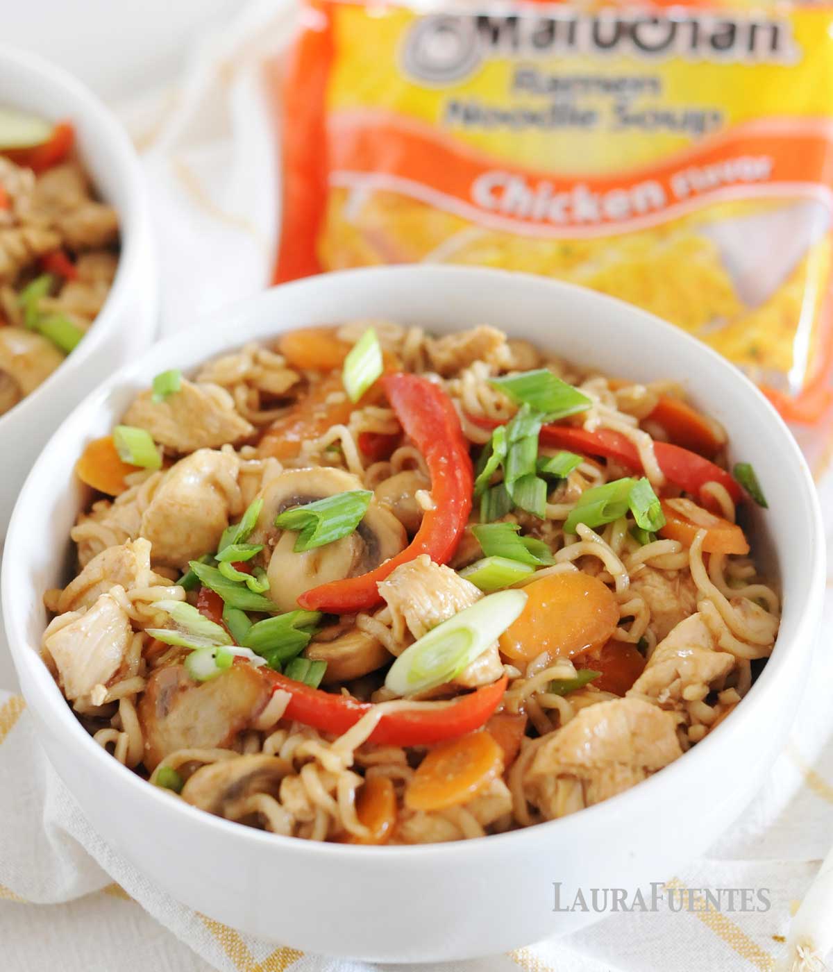 white bowl of ramen noodles, sliced vegetables and chicken with ramen noodles package in backround