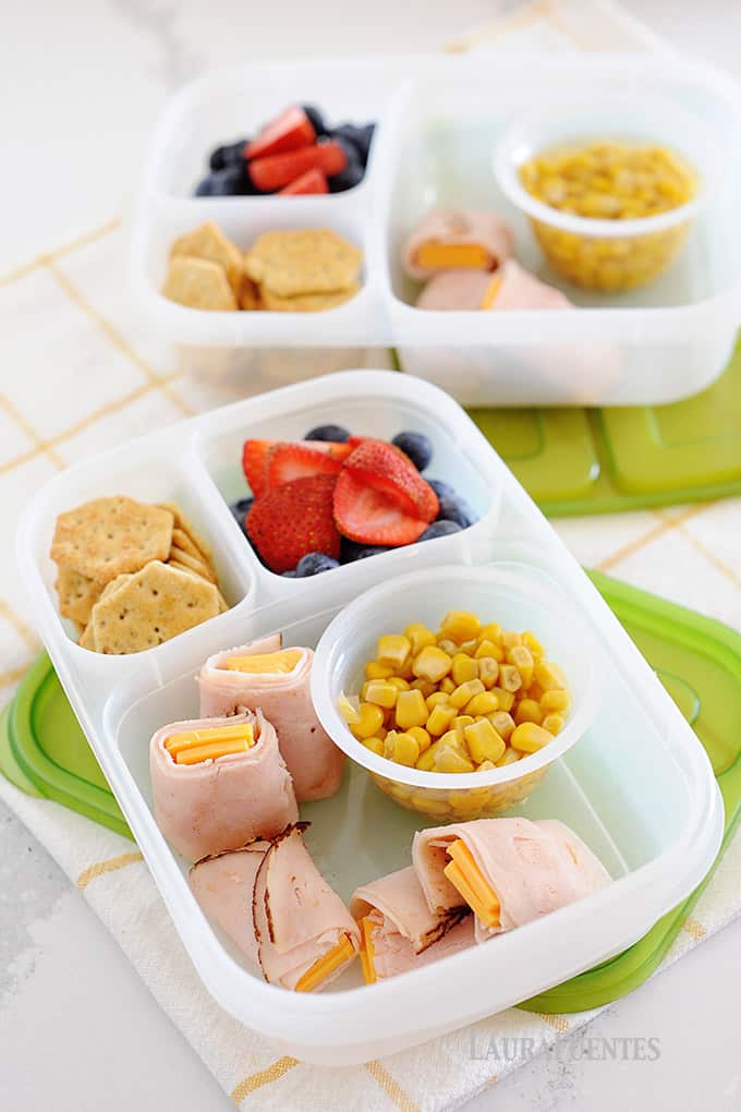 image: two lunch containers filled with crackers, fruit, ham roll ups and corn