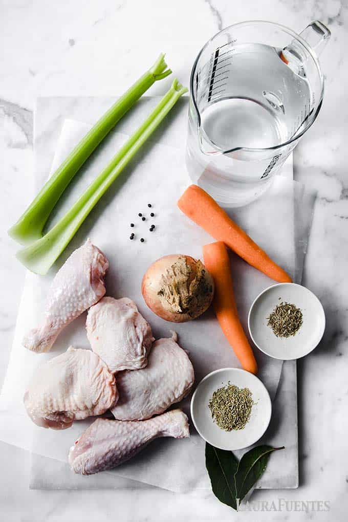 image: Flat lay photo of ingredients for homemade chicken broth. Chicken thighs and legs, carrot sticks, celery sticks, bay leaves, an onion and spices.