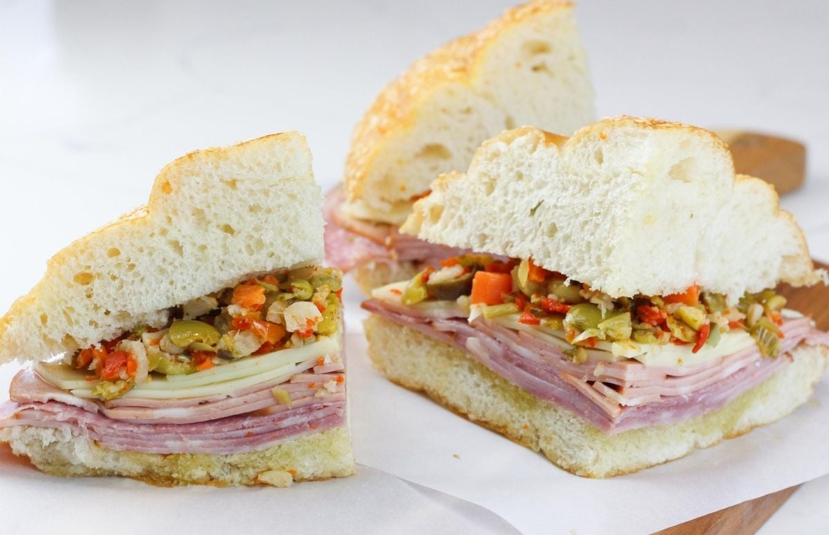 A homemade muffuletta sandwich sliced into quarters on parchment paper on a wooden board