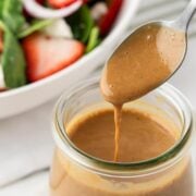 Creamy balsamic vinaigrette drizzling from a spoon into a glass jar with a fresh salad in the background.