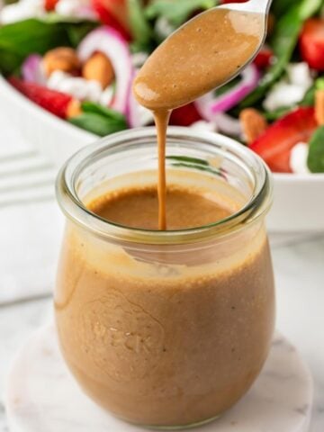Creamy balsamic vinaigrette drizzling from a spoon into a glass jar with a fresh salad in the background.