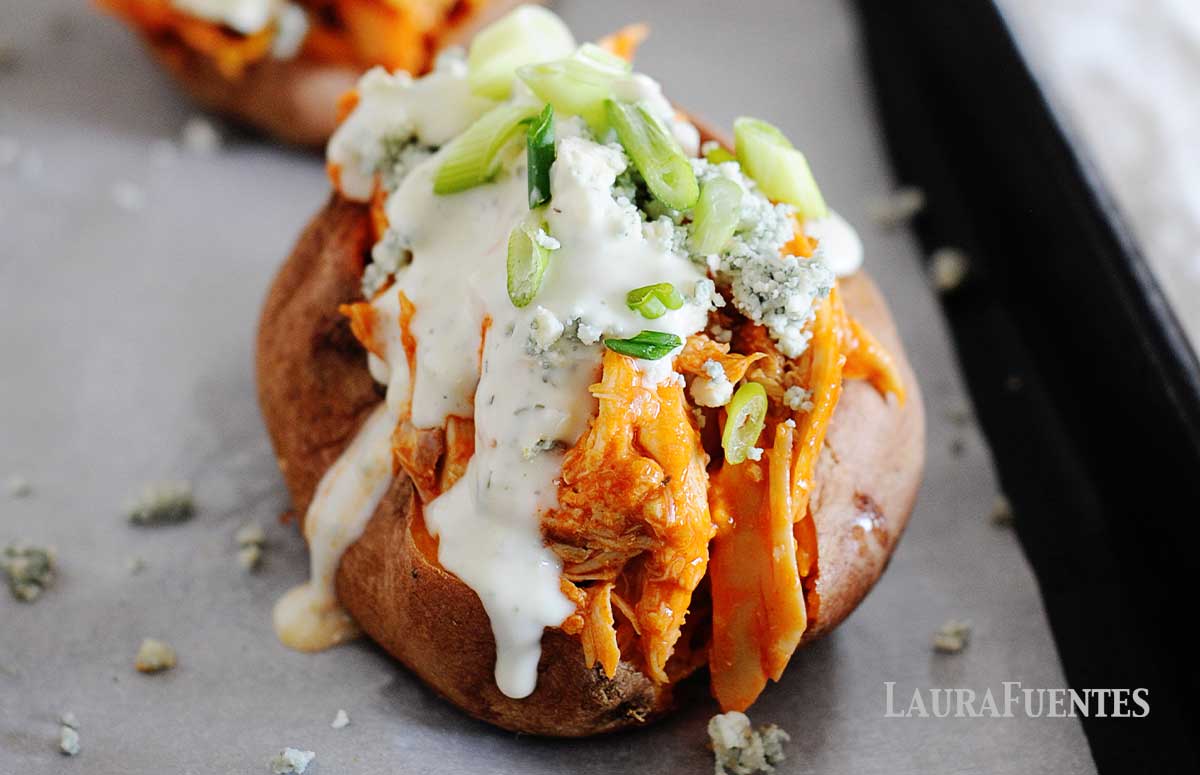 baked sweet potatoes on cooking sheet loaded with buffalo chicken, ranch dressing, green onions and blue cheese crumbles