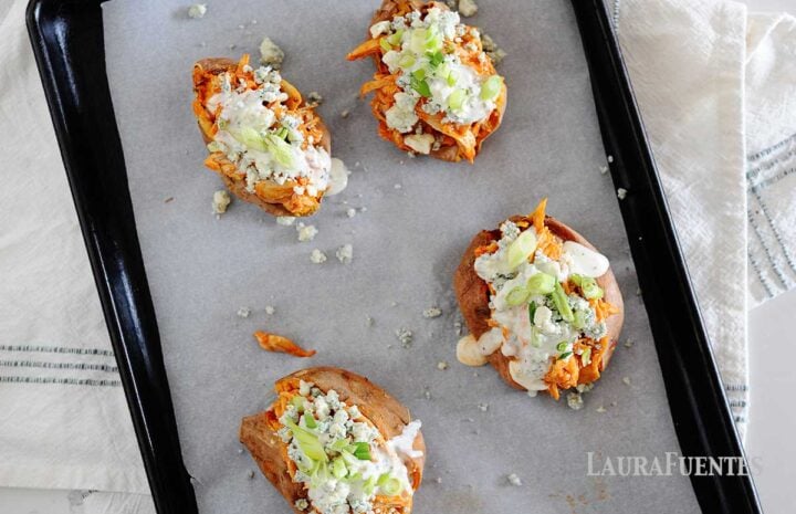 overhead shot of four baked sweet potatoes on cooking sheet loaded with buffalo chicken, ranch dressing, green onions and blue cheese crumbles