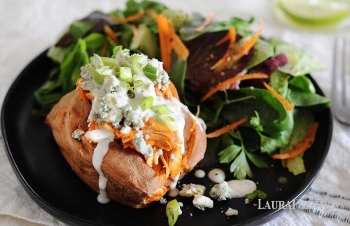 a baked sweet potato with buffalo chicken and blue cheese crumbles on a black plate with salad on the side