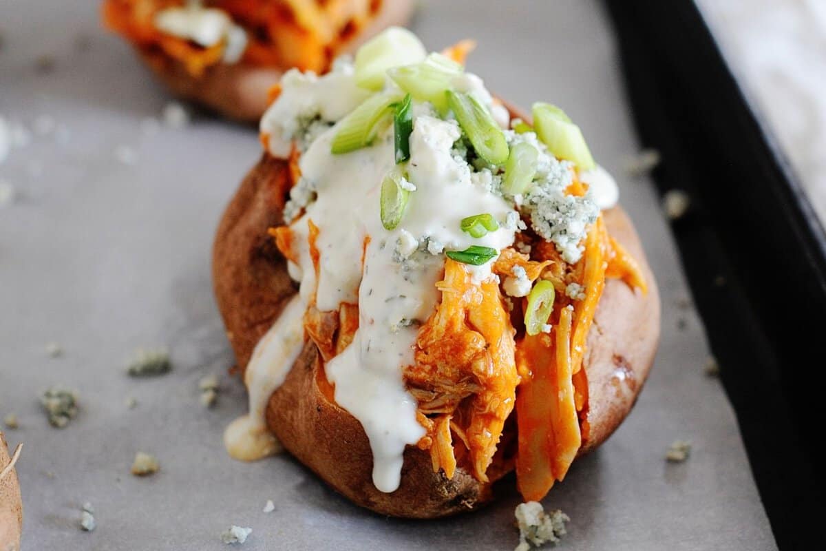baked sweet potatoes on cooking sheet loaded with buffalo chicken, ranch dressing, green onions and blue cheese crumbles