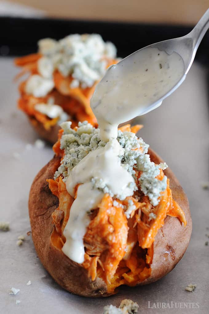 image: two baked sweet potatoes with buffalo chicken and blue cheese crumbles. A spoon of ranch dressing pouring over the top of one potato