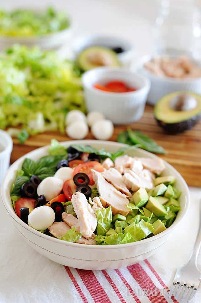 image: Large white bowl of salad with avocado, olives, cheese balls, tomatoes and chicken