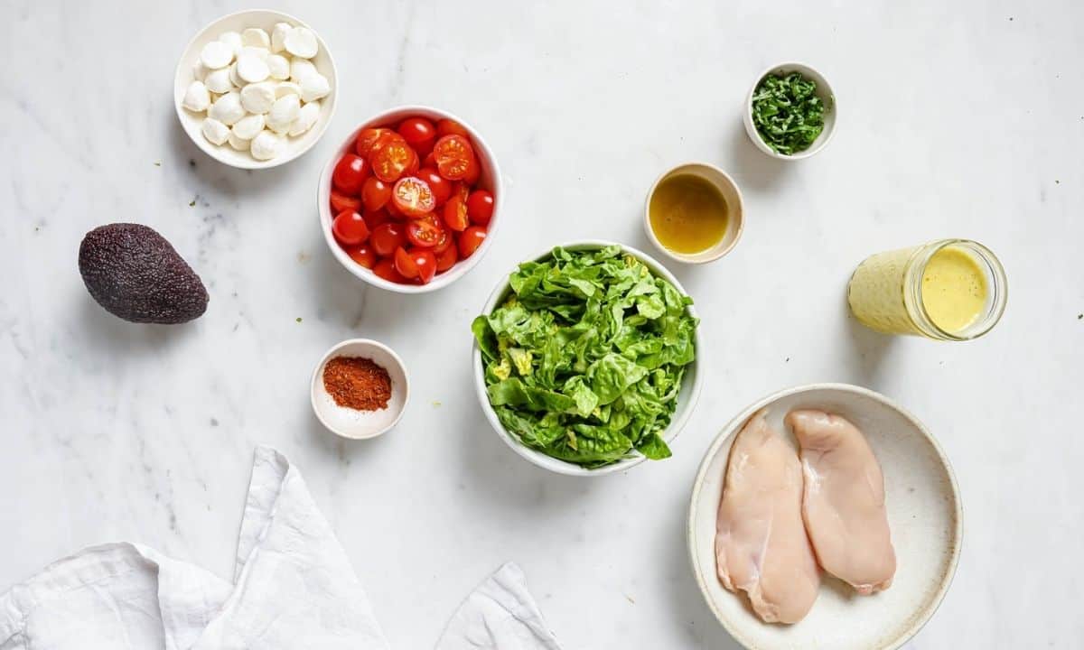 ingredients to make caprese salad: chicken breasts, lettuce, dressing, avocado, cherry tomatoes, bocconcini, basil, oil and a jar of dressing