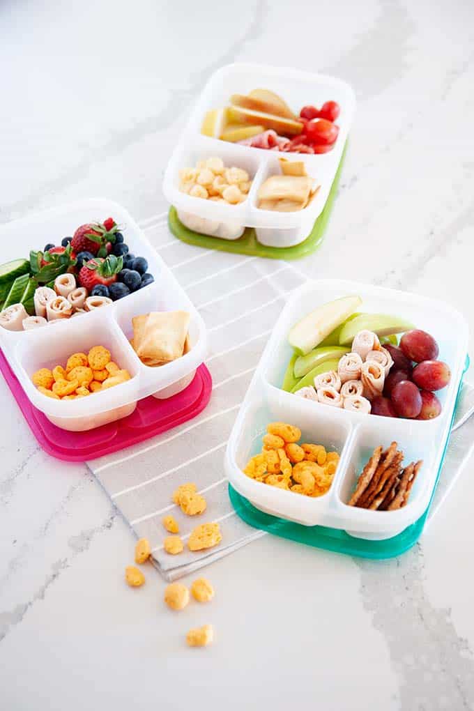 image: three lunchbox containers
filled with high protein snacks like berries, moon cheese, lunch meat and fruit.