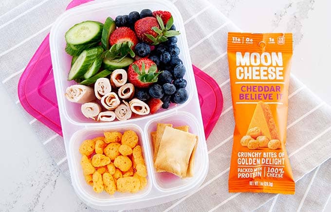 image: overhead shot of snack box filled with pita chips, berries, cucumber slices, turkey rolls and moon cheese snacks. Orange bag of moon cheese snacks next to snack box.