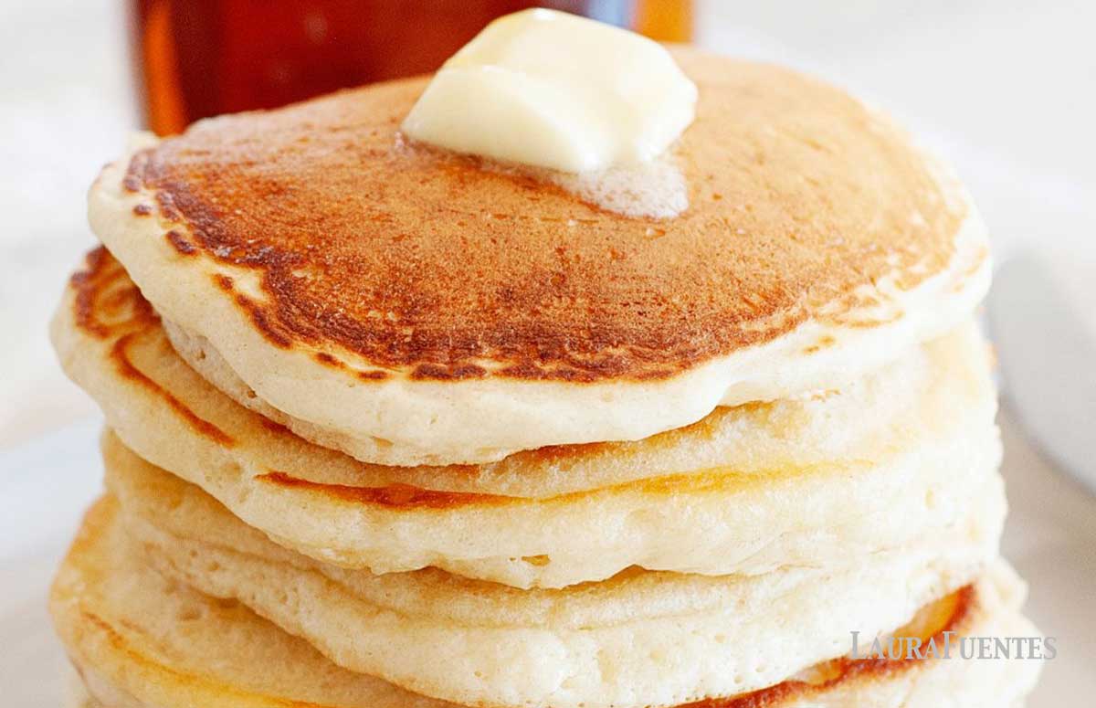 large stack of pancakes topped with a slice of butter. a small bottle of maple syrup behind the stack of pancakes.