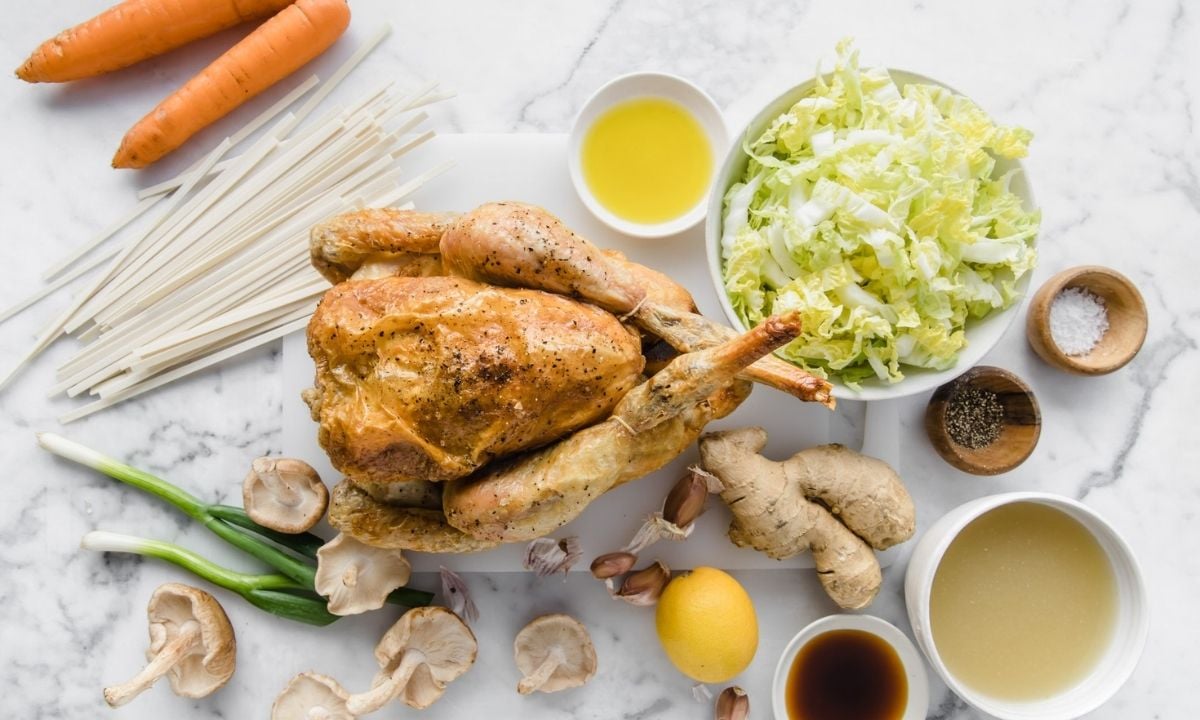 Ingredients for chicken udon soup, including cooked chicken, udon noodles, Napa cabbage, carrots, mushrooms, green onions, ginger, garlic, chicken broth, sesame oil, soy sauce, and lemon arranged on a countertop