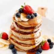 large stack of pancakes, topped with berries, sliced bananas and syrup being poured over the top