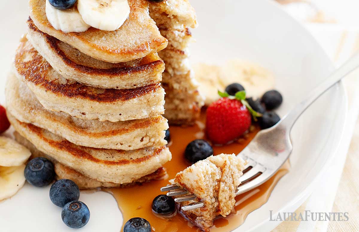 stack of pancakes topped with blueberries, strawberries, sliced bananas. One triangle bite taken out with a fork and placed to the side on the plate