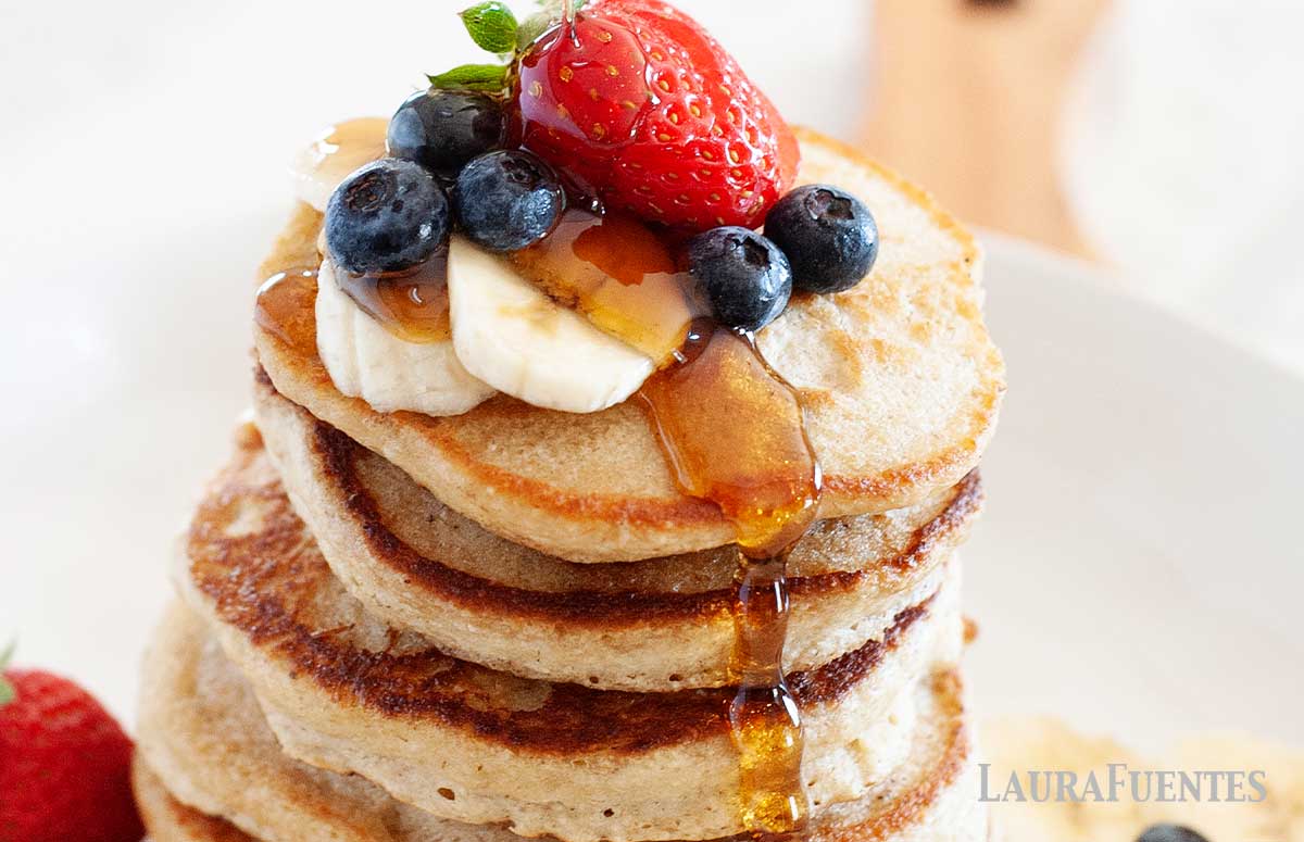stack of six pancakes on a plate topped with berries, bananas and syrup
